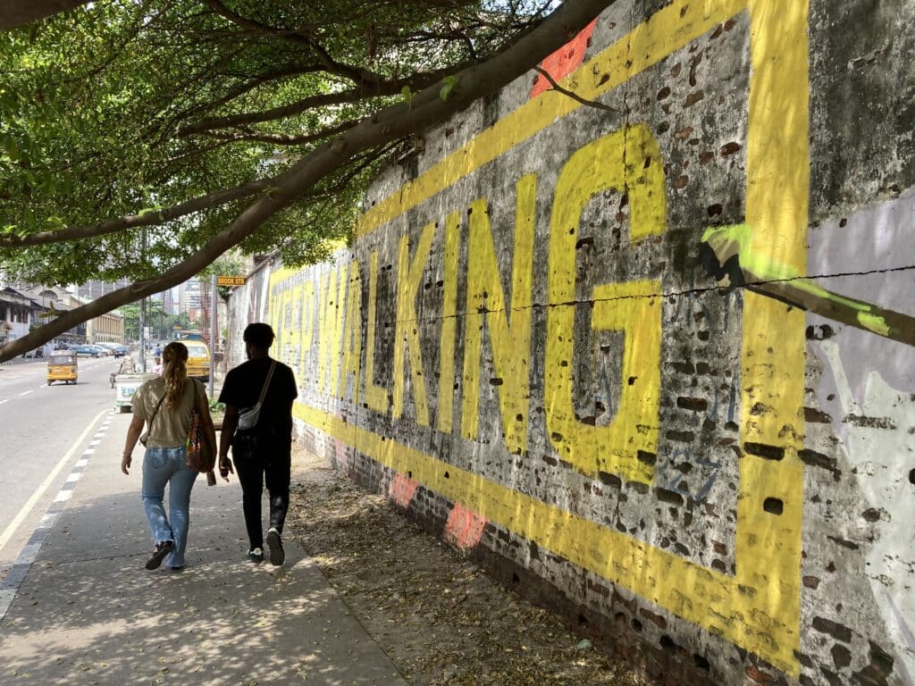 Woman and man walking in Lagos in the shade of a tree, in front of a brick wall that says "walking" in yellow paint.