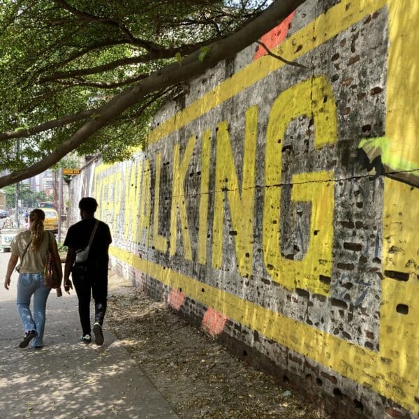Woman and man walking in Lagos in the shade of a tree, in front of a brick wall that says "walking" in yellow paint.