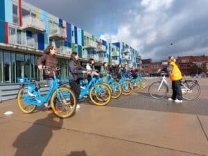 Women standing in a line with bright blue bicycles and yellow tires. A trainer faces them, wearing a yellow jacket.