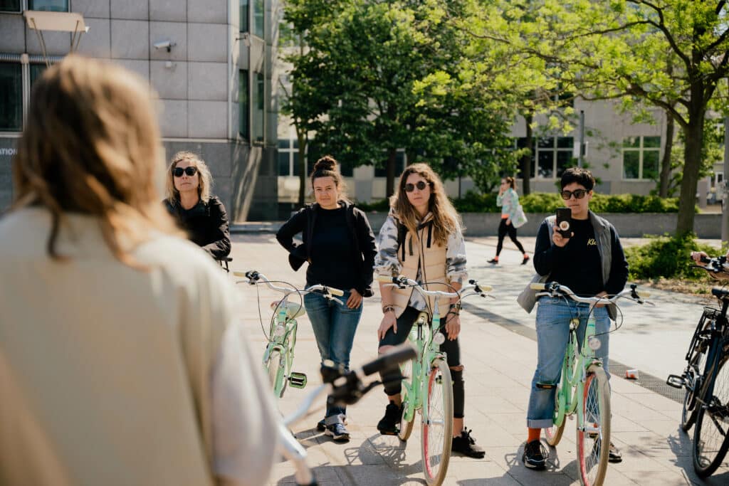Women with bicycles standing in a group, listening to an instructor. The phot is taken over the instructor's shoulder.